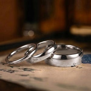 Puede incluir: Tres anillos de metal plateado pulido de diferentes anchos dispuestos en fila. Los anillos tienen una superficie lisa y reflectante con un dise&ntilde;o de banda simple y redondeada. Est&aacute;n colocados sobre una superficie clara con texto oscuro.