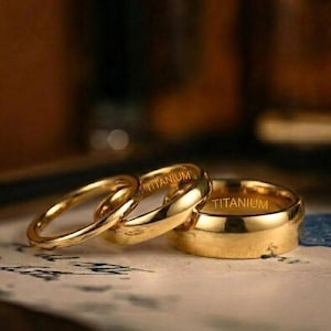 以下が含まれることがあります： 模様のある表面に、細いもの1つと太いもの2つの、3つの金色の指輪が展示されています。「TITANIUM」の文字が太い2つの指輪に刻まれています。背景はぼやけており、暖色系の色合いです。