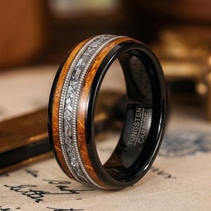 Könnte beinhalten: Ein schwarzer Ring aus Wolframkarbid mit einer mittigen Einlage aus zerkleinerten Meteoritenfragmenten, flankiert von zwei schmalen Bändern aus poliertem Holz. Die Innenseite ist mit dem Text TUNGSTEN CARBIDE in serifenloser Schrift graviert.
