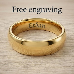 Puede incluir: Un anillo de banda en tono dorado pulido con una superficie exterior lisa y redondeada. El interior del anillo presenta el nombre Ethan grabado en una fuente serif oscura. Sobre el anillo, el texto Free engraving es visible en letras oscuras.
