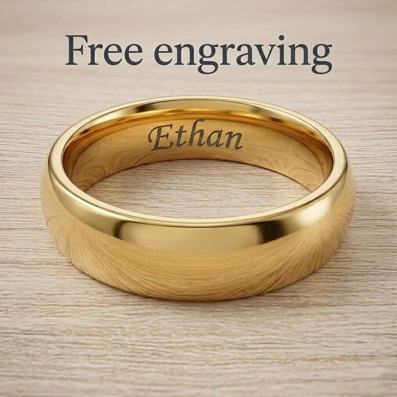 Puede incluir: Un anillo de banda en tono dorado pulido con una superficie exterior lisa y redondeada. El interior del anillo presenta el nombre Ethan grabado en una fuente serif oscura. Sobre el anillo, el texto Free engraving es visible en letras oscuras.