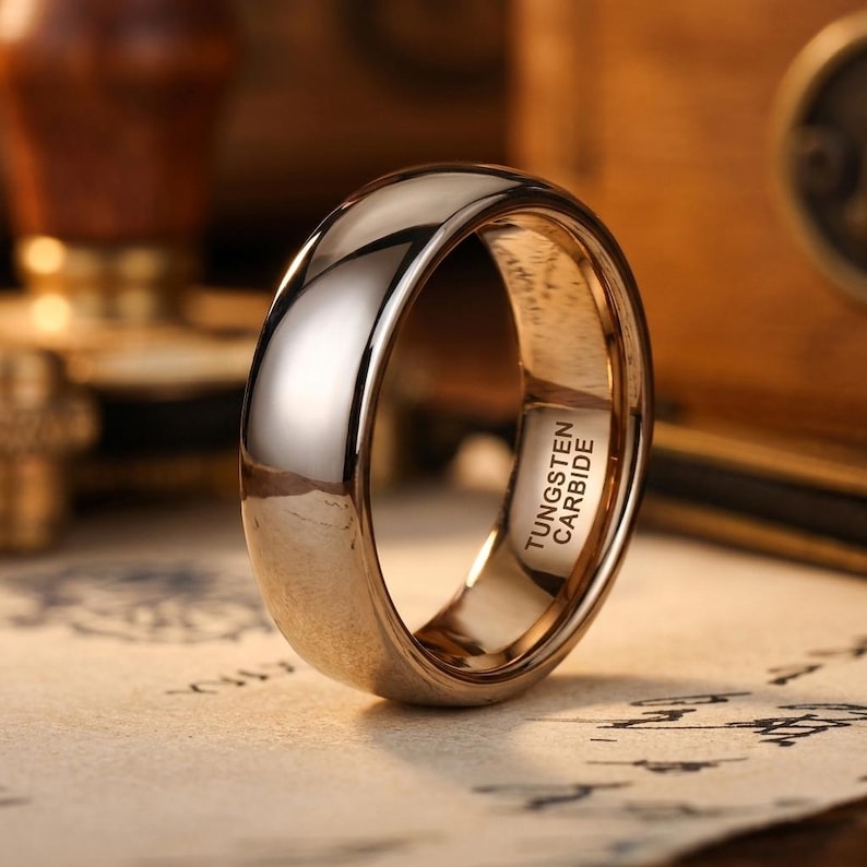 Puede incluir: Un anillo pulido en tono oro rosa con una superficie exterior lisa y abovedada. El interior de la banda est&aacute; claramente grabado con el texto TUNGSTEN CARBIDE, lo que indica la composici&oacute;n material de esta pieza de joyer&iacute;a met&aacute;lica circular.