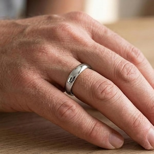Puede incluir: Un anillo de metal plateado pulido con una superficie de banda lisa y redondeada. El anillo presenta una construcci&oacute;n simple y s&oacute;lida con un acabado reflectante de alto brillo que capta la luz, creando un aspecto limpio y uniforme.