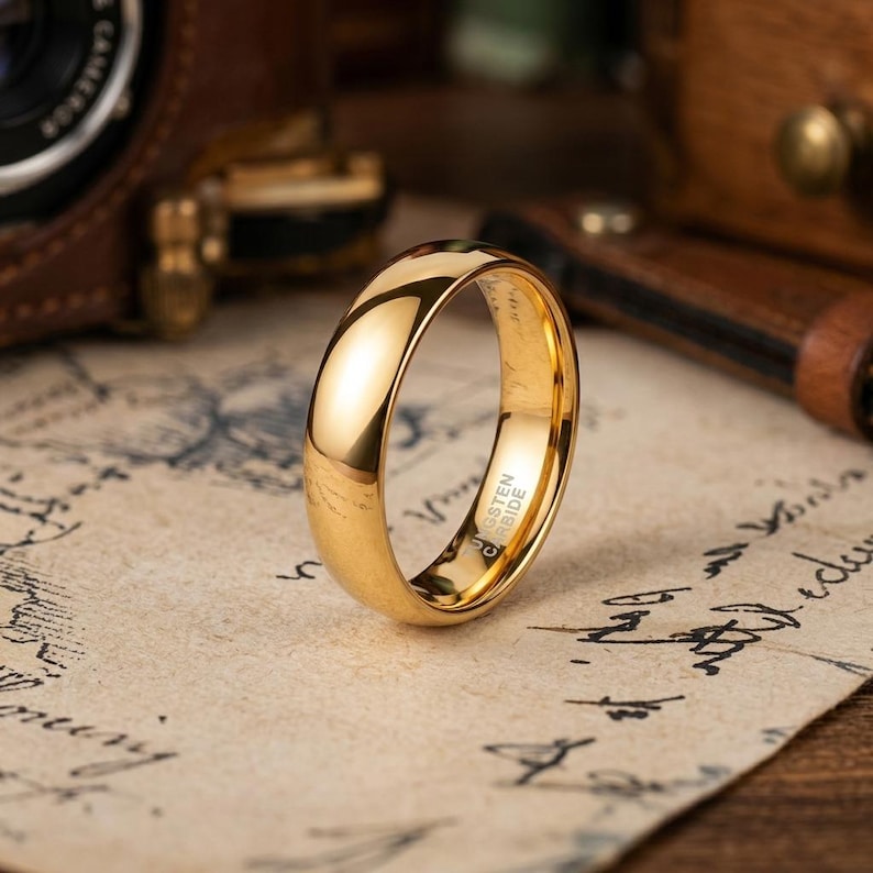 Puede incluir: Un anillo de carburo de tungsteno de color dorado pulido con una superficie exterior lisa y abovedada. El interior de la banda tiene grabado claramente el texto Tungsten Carbide, visible a trav&eacute;s de la abertura central del anillo.
