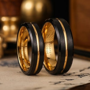 Puede incluir: Dos anillos de boda con exterior negro e interior dorado. Cada anillo tiene una franja dorada en el centro. Las palabras "TUNGSTEN CARBIDE" están impresas en el interior de los anillos.