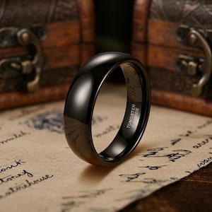 Puede incluir: Un anillo negro pulido con una superficie lisa y abovedada. El interior de la banda est&aacute; claramente grabado con el texto TUNGSTEN CARBIDE en una fuente sans-serif clara. El anillo tiene un acabado de alto brillo que refleja la luz exterior.