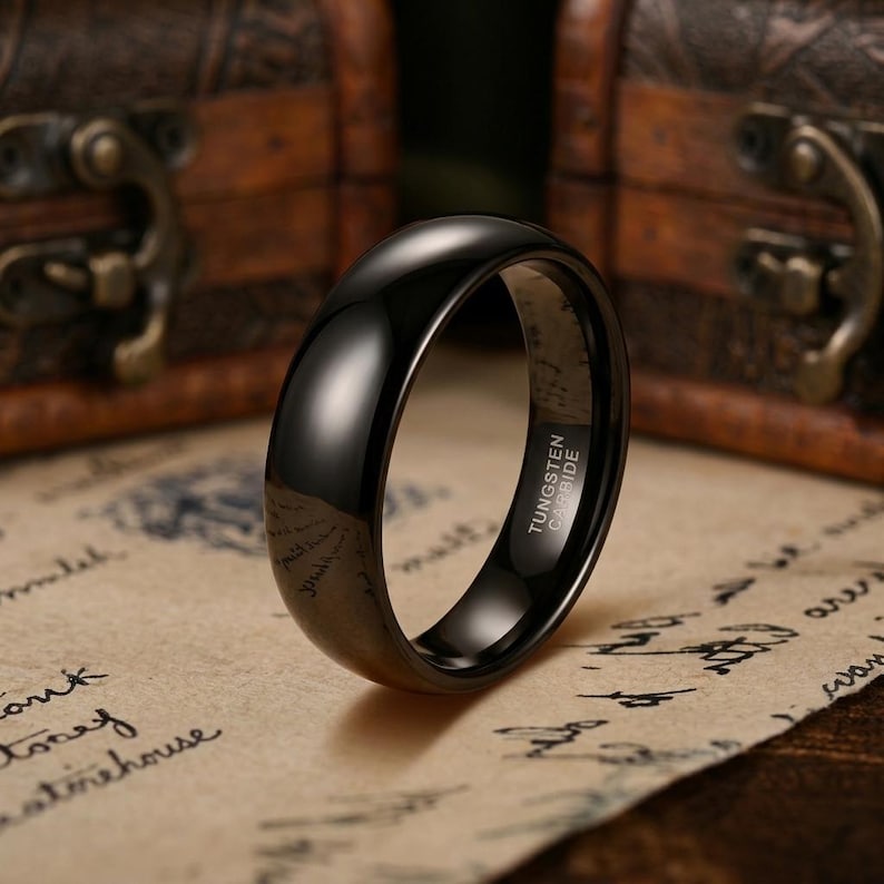 Puede incluir: Un anillo negro pulido con una superficie lisa y abovedada. El interior de la banda est&aacute; claramente grabado con el texto TUNGSTEN CARBIDE en una fuente sans-serif clara. El anillo tiene un acabado de alto brillo que refleja la luz exterior.