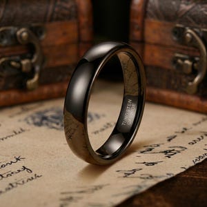 Puede incluir: Un anillo de carburo de tungsteno negro pulido con una superficie lisa y abovedada. El interior de la banda tiene grabado claramente el texto TUNGSTEN CARBIDE, lo que indica la composici&oacute;n del material de esta pieza de joyer&iacute;a circular oscura.