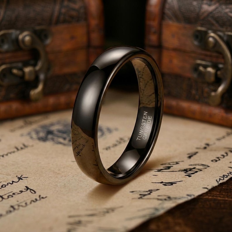 Puede incluir: Un anillo de carburo de tungsteno negro pulido con una superficie lisa y abovedada. El interior de la banda tiene grabado claramente el texto TUNGSTEN CARBIDE, lo que indica la composici&oacute;n del material de esta pieza de joyer&iacute;a circular oscura.