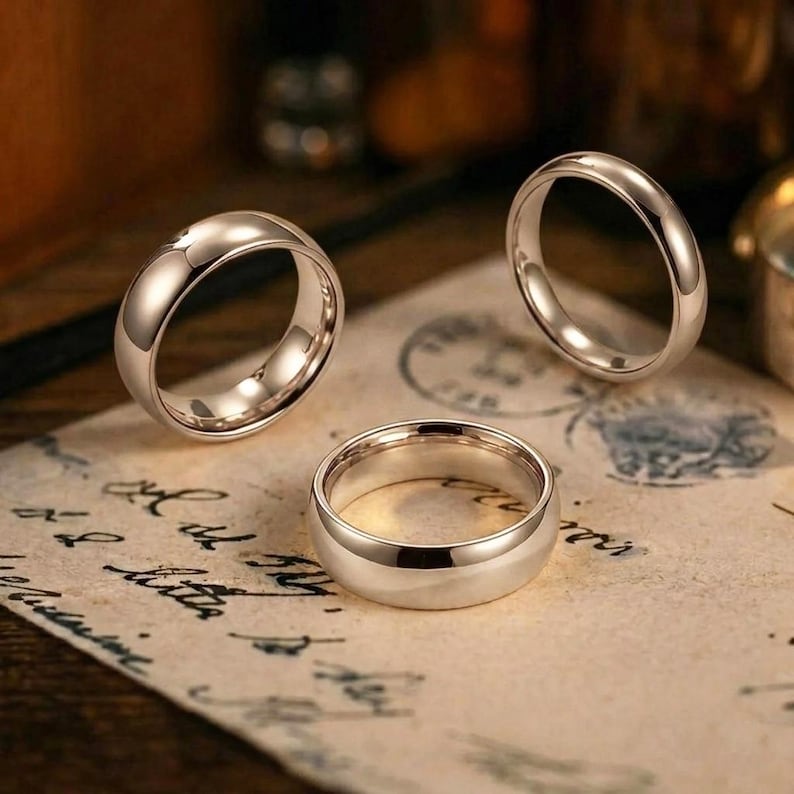 Puede incluir: Tres anillos de metal plateado pulido con un dise&ntilde;o de banda lisa y redondeada. Dos anillos est&aacute;n colocados en posici&oacute;n vertical, mientras que un anillo yace plano de lado. Los anillos presentan un acabado reflectante sin patrones ni piedras.