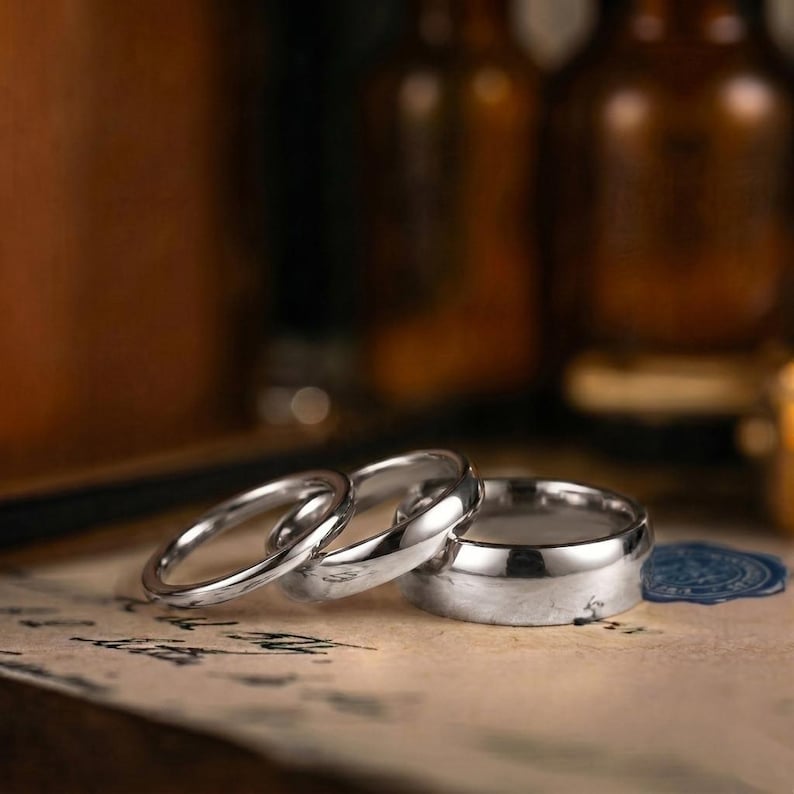 Puede incluir: Tres anillos de metal plateado pulido de diferentes anchos dispuestos en fila. Los anillos tienen una superficie lisa y reflectante con un dise&ntilde;o de banda simple y redondeada. Est&aacute;n colocados sobre una superficie clara con texto oscuro.