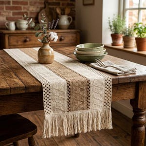 Può includere: Un tavolo rustico in legno è adornato da un runner da tavolo tessuto con una striscia centrale beige affiancata da sezioni color crema con un bordo a frange. Un piccolo vaso con fiori è posto sul runner, accanto a ciotole verdi e un tovagliolo piegato.
