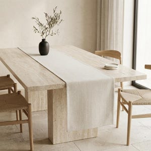 Peut inclure: Une table à manger rectangulaire de couleur claire avec un chemin de table assorti. Un vase noir avec de la verdure est posé sur la table. Des chaises tissées entourent la table, créant un cadre de salle à manger neutre et minimaliste.