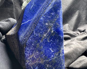 May include: A large, raw piece of deep blue lapis lazuli stone with visible gold-colored pyrite inclusions and white calcite veining. The stone has a polished, angled face and a rough, textured edge, with a pale green crystalline cluster attached.