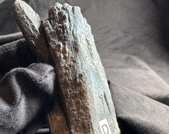 May include: A dark gray, elongated mineral specimen with a rough, crystalline texture and a jagged, pointed top. The surface features natural ridges and indentations, with a small white square sticker labeled with the letter D attached to the base.
