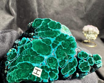 May include: A raw malachite specimen featuring deep green, concentric circular patterns and banded textures. A small white square label with the letter H is attached to the surface of the mineral, which displays a vibrant, polished-like appearance.