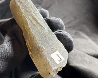 May include: A translucent, pale green, elongated mineral crystal with a rough, textured surface. A small white square sticker with the black letter J is attached to the lower portion of the crystal, which is held against a dark background.