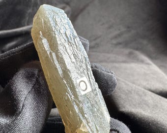 May include: A translucent, pale yellow-green crystal point with a textured, etched surface. A small white rectangular sticker with the letter O printed in black is affixed to the center of the crystal's faceted side.