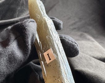 May include: A translucent, pale green, elongated mineral crystal with a jagged, uneven surface texture. A small white rectangular sticker with the black Roman numeral I is attached to the center of the crystal, which features dark inclusions.