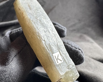 May include: A long, translucent, pale yellow crystal specimen with a hexagonal column shape and a rough, textured surface. A small white square label with the letter K printed in black is attached to the side of the crystal, which has a jagged, semi-transparent base.