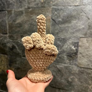 May include: A beige, crocheted sculpture of a hand making the middle finger gesture. The sculpture is held in a person's hand, with a textured, stone-like background.