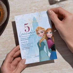Puede incluir: Una tarjeta de invitación azul claro con Elsa y Anna de Frozen. La tarjeta tiene el número 5 en una fuente grande, con texto en español. La invitación es sostenida por una persona.