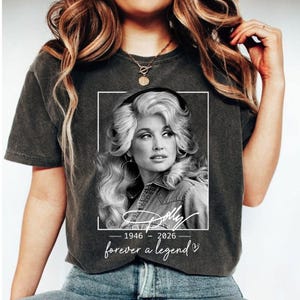 Puede incluir: Una camiseta gris carbón de manga corta con un retrato en blanco y negro de Dolly Parton dentro de un marco rectangular blanco. Debajo del marco, el texto dice Dolly, 1946-2026, y forever a legend con un pequeño icono de corazón.