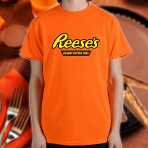 May include: A bright orange short-sleeve t-shirt featuring the yellow and brown Reese's logo printed on the chest. Below the logo, the text reads PEANUT BUTTER CUPS in a brown rectangular graphic that resembles the shape of a candy cup wrapper.