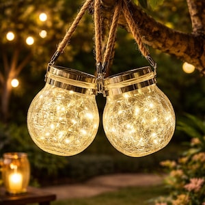 May include: Two round glass lanterns with a crackled texture, each featuring a silver metal lid and a thick jute rope handle. Inside each lantern, warm white string lights glow, creating a bright illumination effect for outdoor spaces.