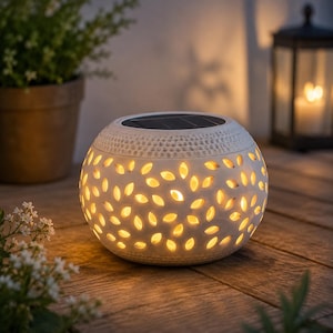 May include: A white, spherical solar-powered lantern with leaf-shaped cutouts, emitting a warm glow. The lantern has a textured top and sits on a wooden surface. A potted plant and a candle lantern are in the background.
