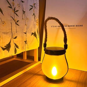 May include: A glowing, yellow lantern with a rope handle. The lantern has a black rim and base. The text on the background reads "IT'S ALL AHE THE MOMENT BEGINS". The lantern is sitting on a wooden surface.