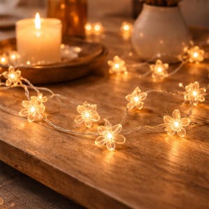 May include: A string of small, clear, flower-shaped LED lights is draped across a wooden surface. The lights are illuminated, casting a warm, inviting glow. A lit candle and other decorative elements are in the background, creating a cozy ambiance.