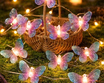 Könnte beinhalten: Eine LED-Lichterkette mit durchscheinenden, schmetterlingsförmigen Abdeckungen in Violett- und Blautönen. Jeder Schmetterling enthält in der Mitte ein kleines leuchtendes Licht. Die Lichterkette ist um und in einem kleinen geflochtenen Korb drapiert.
