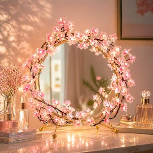 May include: A round mirror with a gold frame, adorned with pink cherry blossom flowers and small lights. The mirror is on a gold stand, and the scene includes perfume bottles and a vase of pink flowers, creating a soft, romantic ambiance.