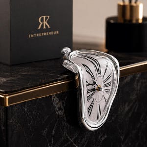 Puede incluir: Un reloj plateado y derretido con una esfera blanca y números romanos negros. El reloj descansa sobre una superficie de mármol negro. Una caja negra con la palabra "ENTREPRENEUR" está en el fondo.