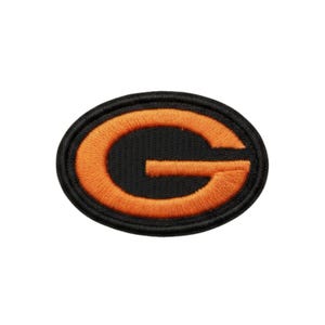 May include: An embroidered patch with a black oval border and an orange "G" in the centre. The letter is stylized and filled with orange thread, contrasting against the black background.