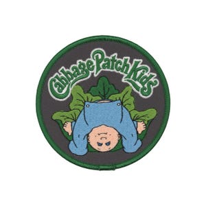 May include: Embroidered patch featuring a Cabbage Patch Kids design. The patch is circular with a dark gray background and a green border. The design includes a baby in a blue outfit, green leaves, and the words "Cabbage Patch Kids" in green.