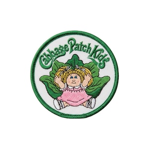 May include: Embroidered patch featuring a Cabbage Patch Kids doll. The design includes the words "Cabbage Patch Kids" in green script above the doll. The doll is wearing a pink dress and has blonde pigtails. The patch has a white background with a green border.