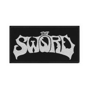 May include: Black rectangular patch with the white text "THE SWORD" in a stylized, retro font. The patch has a black border and is likely made of fabric, suitable for sewing or ironing onto clothing or accessories.