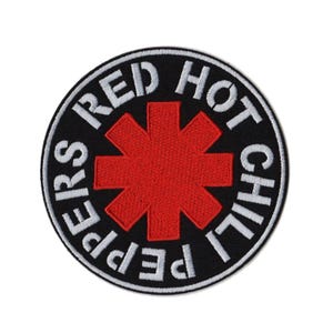 Peut inclure: Un écusson rond noir avec l'inscription blanche "RED HOT CHILI PEPPERS". Le centre présente un symbole rouge en forme d'astérisque. L'écusson est probablement destiné aux vêtements ou aux accessoires.