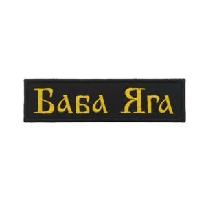 Puede incluir: Parche rectangular negro con texto cirílico bordado en amarillo que dice "Baba Yaga". El parche tiene un borde negro y probablemente está hecho de tela. El texto está en negrita, con una fuente estilizada.