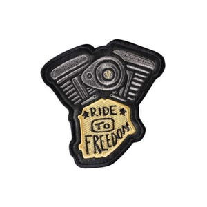 以下が含まれることがあります： 黒とグレーの刺繍パッチ。バイクのエンジンデザインが特徴です。「RIDE TO FREEDOM」の文字がスタイリッシュなフォントで、ゴールドとブラックの配色でデザインされています。ジャケットやバッグなどに使用される可能性があります。