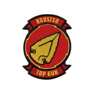 Pode incluir: Patch bordado com um círculo vermelho e um design de seta amarela. O patch tem uma borda preta e faixas vermelhas na parte superior e inferior. A faixa superior diz "ROOSTER", a inferior "TOP GUN".