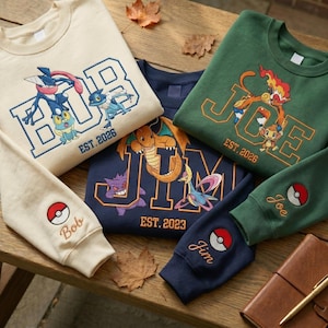 Könnte beinhalten: Drei Rundhals-Sweatshirts in Creme, Marineblau und Waldgrün. Jedes zeigt große gestickte Blockbuchstaben mit Namen wie BOB, JIM und JOE, begleitet von Zeichentrickfiguren und dem Text EST. 2023 oder 2026. Die Ärmel sind mit Pokéball-Symbolen versehen.