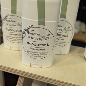 May include: A group of white deodorant sticks with green stripes and labels. The labels display "meadow & bloom tallow deodorant lemongrass." The sticks are presented on a light wooden shelf.