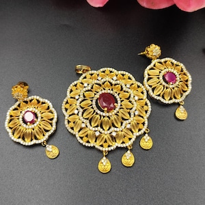 Puede incluir: Un conjunto de joyas de tono dorado con un colgante y pendientes a juego. El colgante y los pendientes tienen un diseño floral con una gema roja central y están adornados con pequeñas piedras transparentes. Los pendientes tienen un colgante en forma de lágrima.