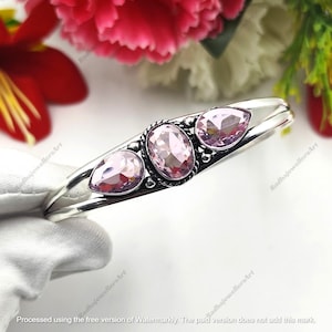 May include: A silver cuff bracelet featuring three faceted light pink gemstones. The center stone is oval-shaped, flanked by two pear-shaped stones. The metal setting has a twisted rope texture around the center stone and small silver beads.