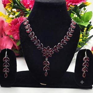 Puede incluir: Un conjunto de collar y pendientes colgantes en tono plateado con piedras de color rojo oscuro talladas en marquise, dispuestas en un patrón de hojas. El collar incluye un motivo floral central con un colgante en forma de hoja.