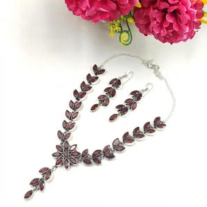 May include: A silver-toned necklace and matching earring set featuring marquise-cut dark red stones arranged in a leaf-like pattern. The necklace includes a central flower-shaped cluster of stones, and both pieces are connected by thin silver chains.