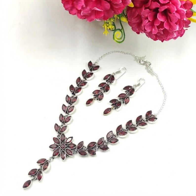 May include: A silver-toned necklace and matching earring set featuring marquise-cut dark red stones arranged in a leaf-like pattern. The necklace includes a central flower-shaped cluster of stones, and both pieces are connected by thin silver chains.